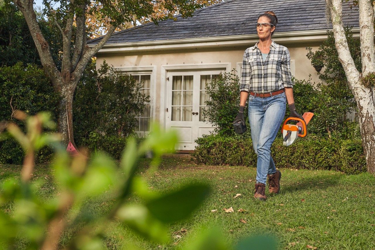 Una mujer camina sobre el césped delante de una casa con un cortasetos STIHL