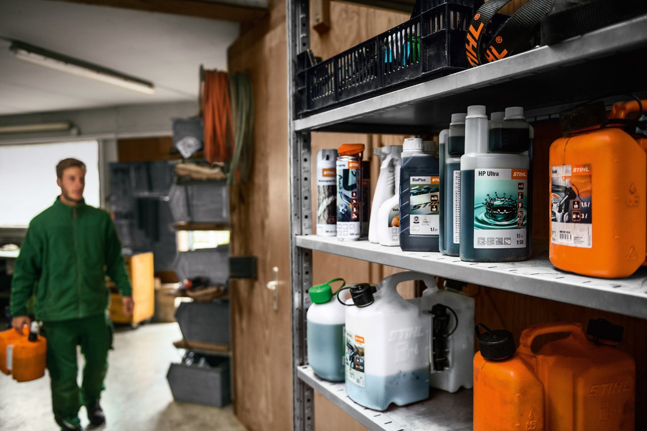 Varios materiales consumibles STIHL en una estantería, un hombre con bidón al fondo para el transporte de mercancías peligrosas