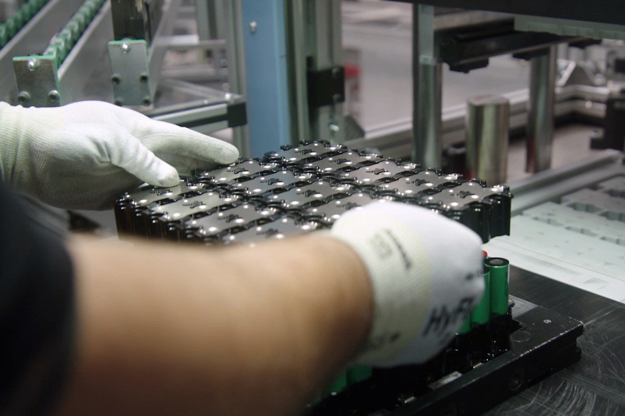 Un hombre con guantes de trabajo coloca el soporte superior de celdas, provisto de placas de unión de celdas, en el soporte inferior para unir las celdas de la batería